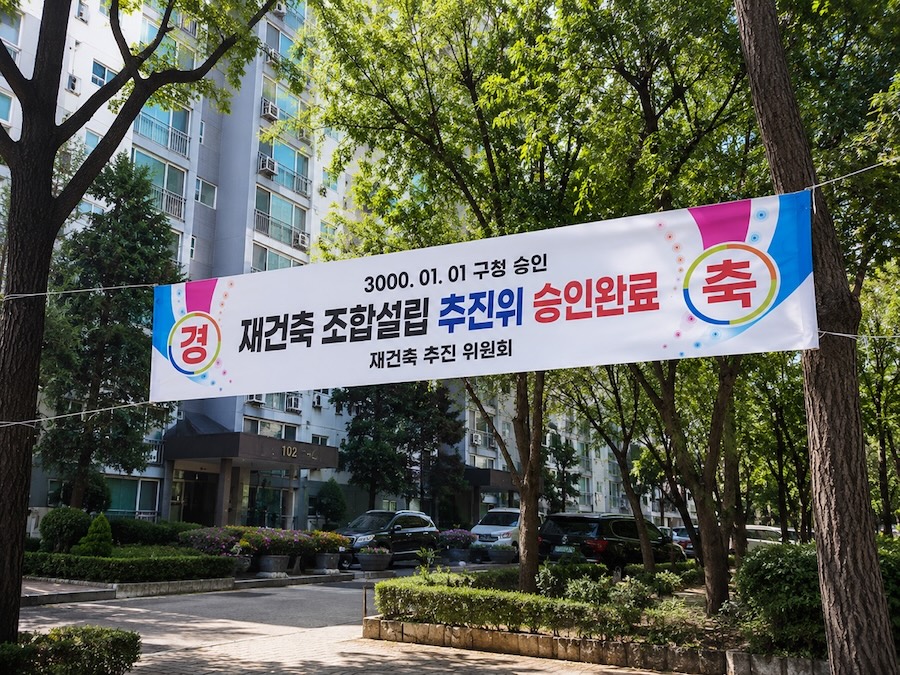 아파트 단지 입구 나무 사이에 걸린 '재건축 조합설립 추진위 승인완료 — 재건축 추진위원회' 축하 현수막.