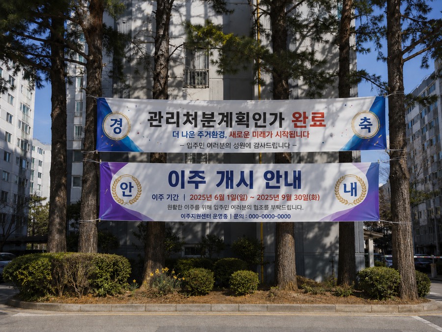 아파트 단지 입구에 위아래로 걸린 현수막 두 개. 위는 '관리처분계획인가 완료' 축하 현수막, 아래는 이주 기간을 안내하는 '이주 개시 안내' 현수막.